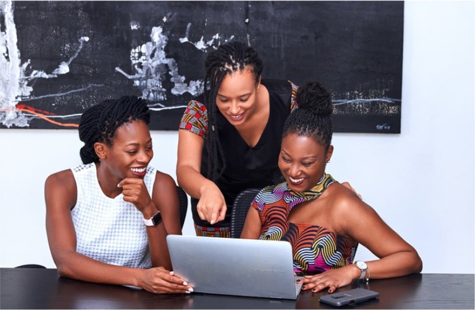 Three women lean over a laptop together, sharing a smile.