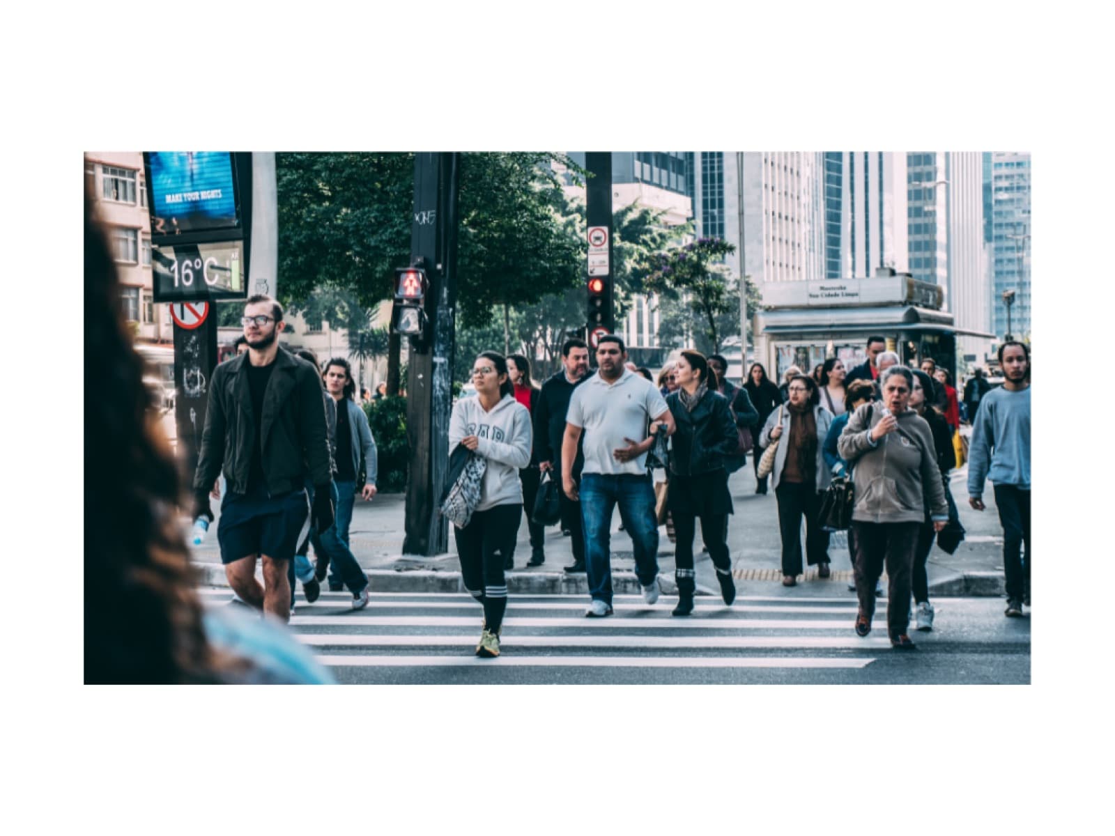 Commuters cross a wide city avenue past storefronts and trees.